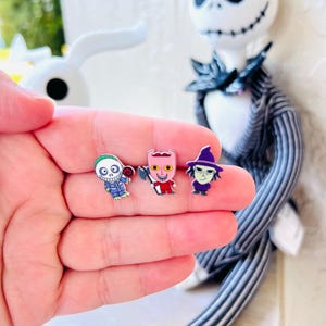 May include: Three enamel pins featuring characters from The Nightmare Before Christmas. The pins depict characters in vibrant colors: a blue and white skeleton, a red and white creature, and a purple witch. A Jack Skellington doll is in the background.