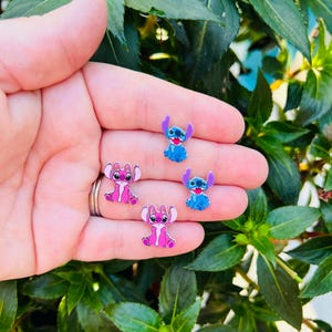 Może przedstawiać: Kolekcja małych, kolorowych zawieszek z postacią Stitch w kolorze niebieskim i r&oacute;żowym. Zawieszki umieszczone są na tle zielonych liści. Zawieszki są prawdopodobnie przeznaczone do biżuterii lub rękodzieła.