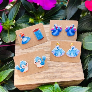 May include: A collection of four pairs of earrings displayed on small brown cards. The earrings feature cartoon designs of a princess, a spool of thread, a blue bird, and a princess in a blue dress. The earrings are set against a backdrop of green leaves and pink flowers.