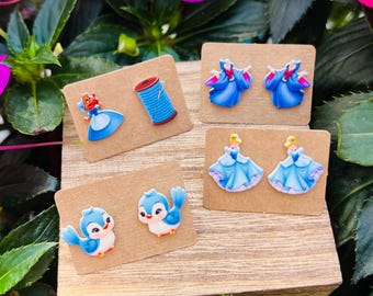 May include: A collection of four pairs of earrings displayed on small brown cards. The earrings feature cartoon designs of a princess, a spool of thread, a blue bird, and a princess in a blue dress. The earrings are set against a backdrop of green leaves and pink flowers.