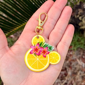 Puede incluir: Un llavero de rodaja de lim&oacute;n amarillo con un cierre dorado. El llavero presenta un dise&ntilde;o de rodajas de lim&oacute;n, flores rosas y hojas verdes. El llavero se sostiene en una mano.