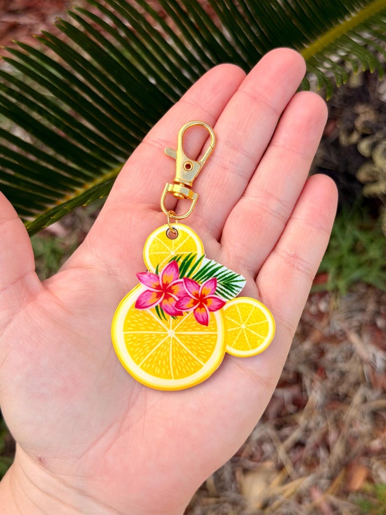 Puede incluir: Un llavero de rodaja de lim&oacute;n amarillo con un cierre dorado. El llavero presenta un dise&ntilde;o de rodajas de lim&oacute;n, flores rosas y hojas verdes. El llavero se sostiene en una mano.
