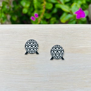 May include: A pair of black and white geometric sphere earrings. Each earring features a detailed pattern of triangles and a black base. The earrings are set against a light wood grain surface with a blurred green and purple background.