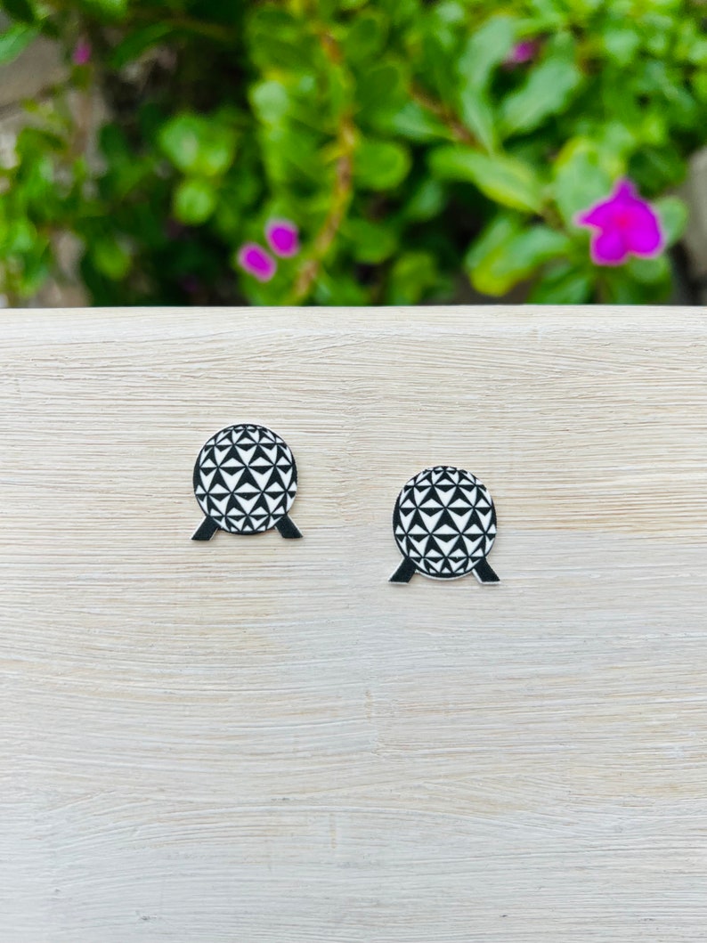 May include: A pair of black and white geometric sphere earrings. Each earring features a detailed pattern of triangles and a black base. The earrings are set against a light wood grain surface with a blurred green and purple background.