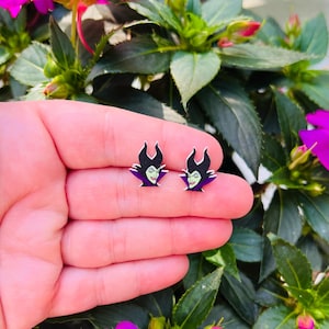 Puede incluir: Un par de pendientes con una imagen de dibujos animados de Mal&eacute;fica, de la pel&iacute;cula de Disney La Bella Durmiente, en morado y verde.