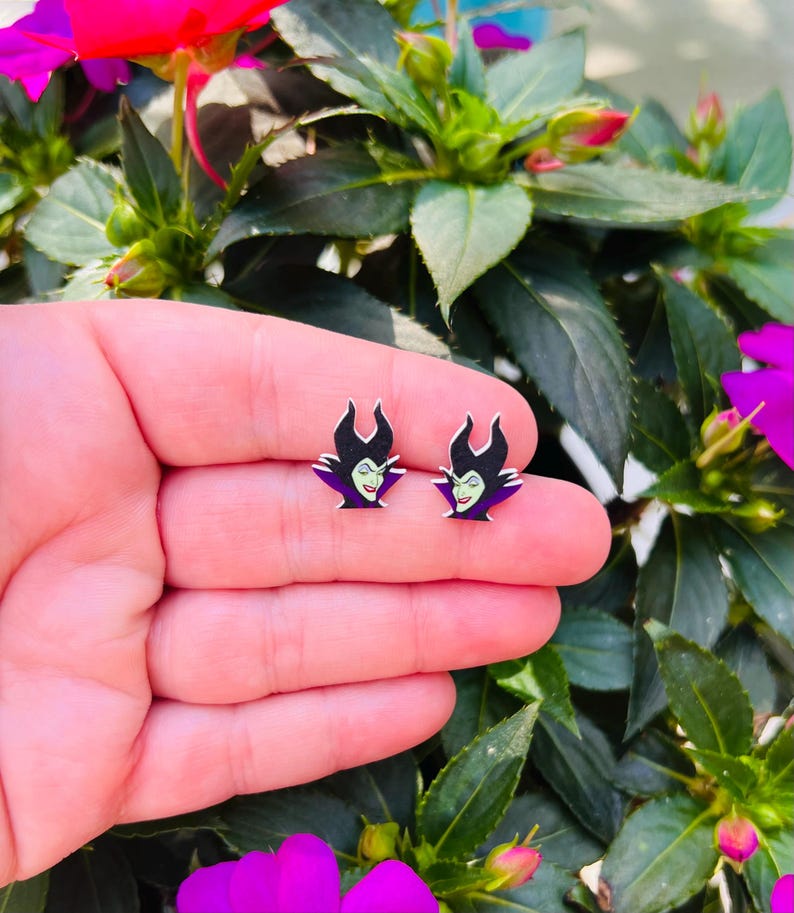 Puede incluir: Un par de pendientes con una imagen de dibujos animados de Mal&eacute;fica, de la pel&iacute;cula de Disney La Bella Durmiente, en morado y verde.