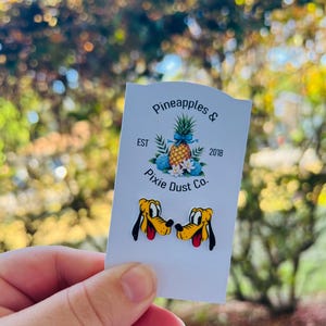 May include: A white card with the text "Pineapples & Pixie Dust Co. EST 2018" and a cartoon image of two yellow and brown Pluto dog characters.