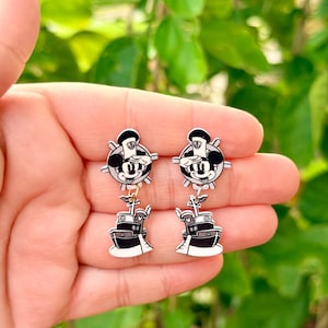 May include: A pair of black and white dangle earrings featuring a cartoon mouse character wearing a sailor hat above a black and white boat. The earrings are held in a hand against a blurred green background.