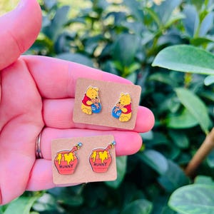 May include: Two pairs of Winnie the Pooh themed earrings. One pair features Winnie the Pooh holding a honey pot, the other pair features a honey pot with the word "Hunny" on it.
