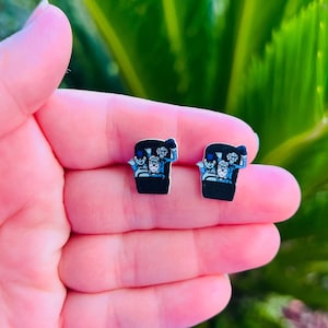 May include: A pair of black and blue stud earrings. Each earring features a cartoon image of a group of people in a vehicle, with arms raised in the air. The earrings are set against a blurred green background.