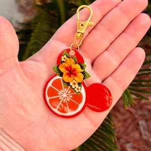 Może przedstawiać: Czerwony brelok w kształcie Myszki Miki ze złotym zapięciem. Brelok przedstawia plaster pomarańczy i kwiatowy wz&oacute;r. Plaster pomarańczy jest biało-pomarańczowy. Kwiat jest ż&oacute;łto-pomarańczowy z zielonymi liśćmi.