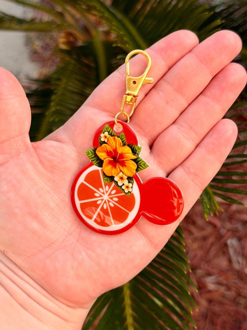 Może przedstawiać: Czerwony brelok w kształcie Myszki Miki ze złotym zapięciem. Brelok przedstawia plaster pomarańczy i kwiatowy wz&oacute;r. Plaster pomarańczy jest biało-pomarańczowy. Kwiat jest ż&oacute;łto-pomarańczowy z zielonymi liśćmi.