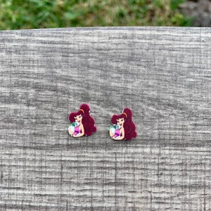 May include: Two small, maroon-haired cartoon character earrings. Each earring features a woman in a purple dress holding a white flower. The earrings are on a gray wooden surface.