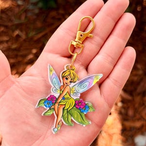 May include: A colorful keychain featuring a fairy with yellow hair, a green dress, and iridescent wings. The fairy is seated on green leaves with pink and blue flowers. The keychain has a gold-colored clasp.