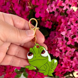 May include: A green enamel keychain featuring a cartoon frog with red eyes and a gold-colored clasp. The frog has a whimsical design and is set against a backdrop of vibrant pink flowers.