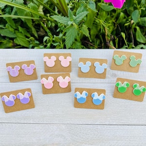 May include: A collection of colorful Mickey Mouse-shaped stud earrings. Each pair is mounted on a small, rectangular cardboard backing. The earrings feature various colors, including pink, blue, green, and purple, with striped patterns.