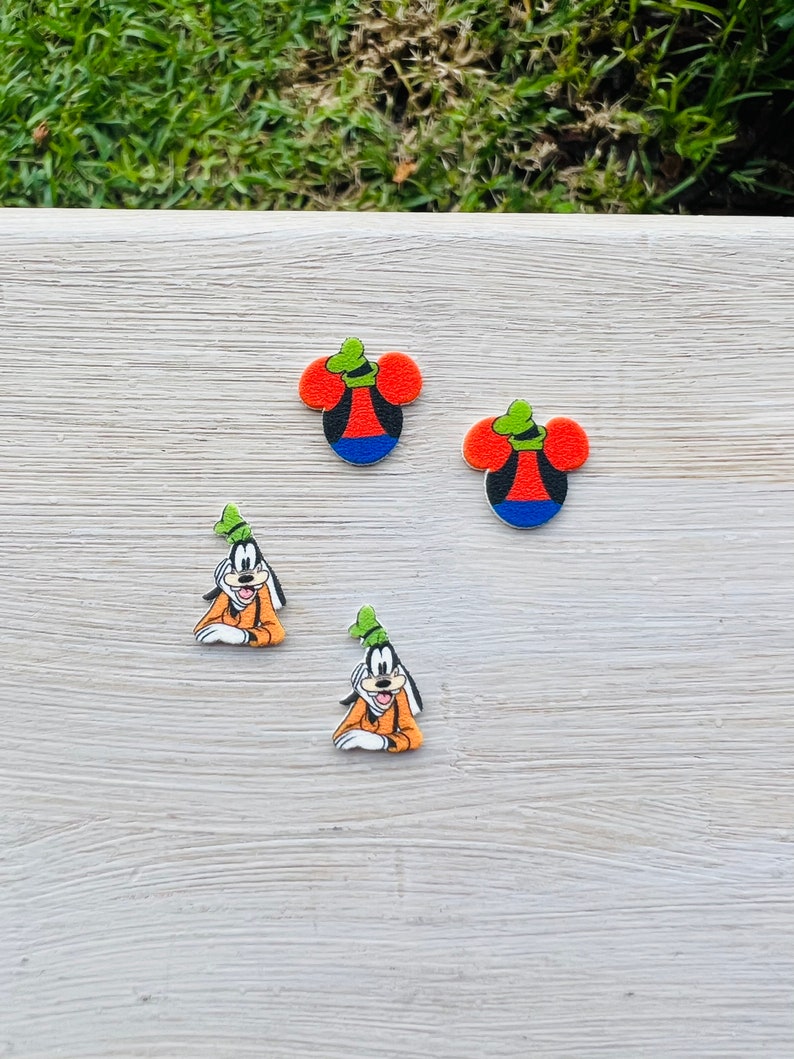 May include: Four colorful cartoon character charms. Two charms are shaped like Mickey Mouse's head, with red ears, a black face, a blue bottom, and a green hat. Two charms depict Goofy in an orange outfit and a green hat.