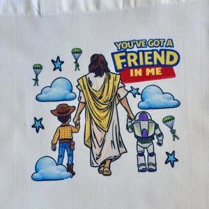 Pu&ograve; includere: Borsa tote bianca con una grafica colorata con la scritta "YOU'VE GOT A FRIEND IN ME". Il design include personaggi dei cartoni animati, nuvole, stelle e paracadute. La borsa ha due manici bianchi.