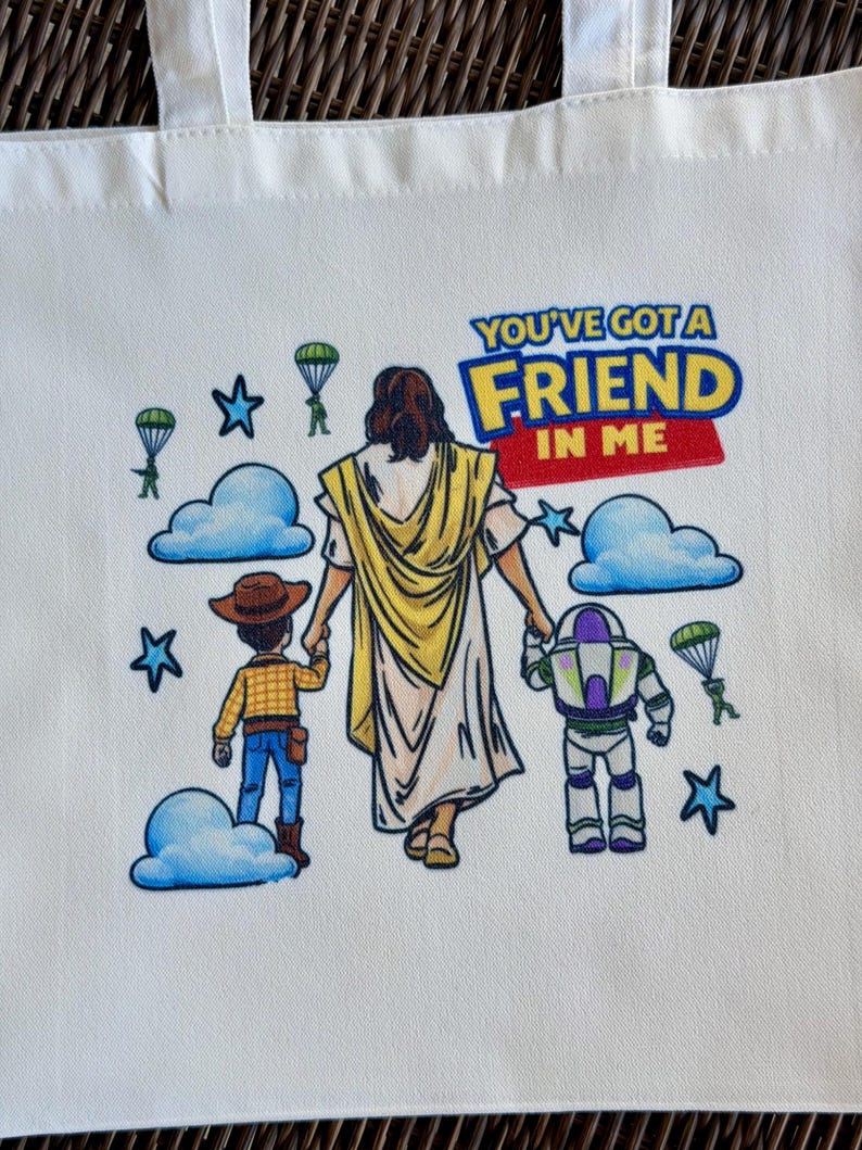 Pu&ograve; includere: Borsa tote bianca con una grafica colorata con la scritta "YOU'VE GOT A FRIEND IN ME". Il design include personaggi dei cartoni animati, nuvole, stelle e paracadute. La borsa ha due manici bianchi.