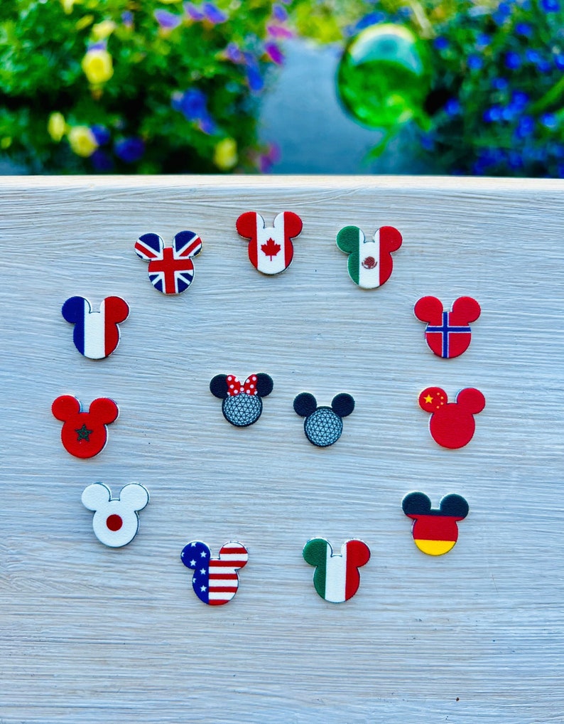 May include: A selection of Mickey Mouse-shaped charms, each displaying a different national flag, such as the Union Jack, Canadian, Mexican, French, Norwegian, Chinese, Japanese, and American flags. Some charms feature a golf ball design. The charms are on a light wood surface.