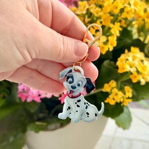 May include: A Dalmatian puppy keychain with a gold-colored clasp. The keychain features a white puppy with black spots, a red collar, and black ears. The background is blurred with pink and yellow flowers.