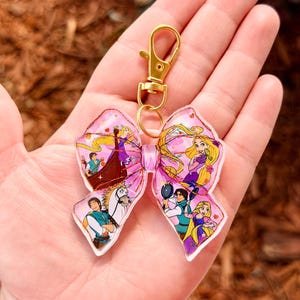 May include: A pink bow-shaped keychain featuring cartoon characters. The bow has a gold-colored clasp and ring. The bow's design includes images of a princess with long blonde hair and other characters from a fairytale.