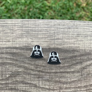 May include: Two black and white Darth Vader helmet-shaped earrings. The earrings are on a gray wooden surface with a blurred green background. The earrings are a Star Wars-themed accessory.