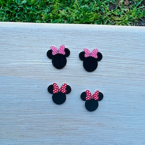 May include: Four black mouse-shaped embellishments with red and pink polka dot bows. The embellishments are arranged on a white wooden surface with green grass in the background. The bows are in different sizes.