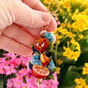 May include: A colorful keychain featuring a cartoon bird character. The bird is perched on a slice of orange, surrounded by blue flowers and green leaves. The keychain has a gold-colored clasp.