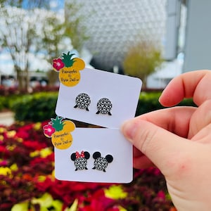 May include: Two pairs of stud earrings on white cards are held up in front of a blurred background. One pair features a geometric design, and the other has a Minnie Mouse design with a red bow. The cards have a pineapple logo with text.
