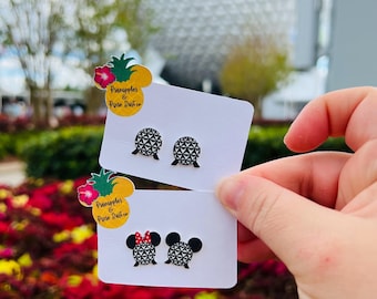 May include: Two pairs of stud earrings on white cards are held up in front of a blurred background. One pair features a geometric design, and the other has a Minnie Mouse design with a red bow. The cards have a pineapple logo with text.