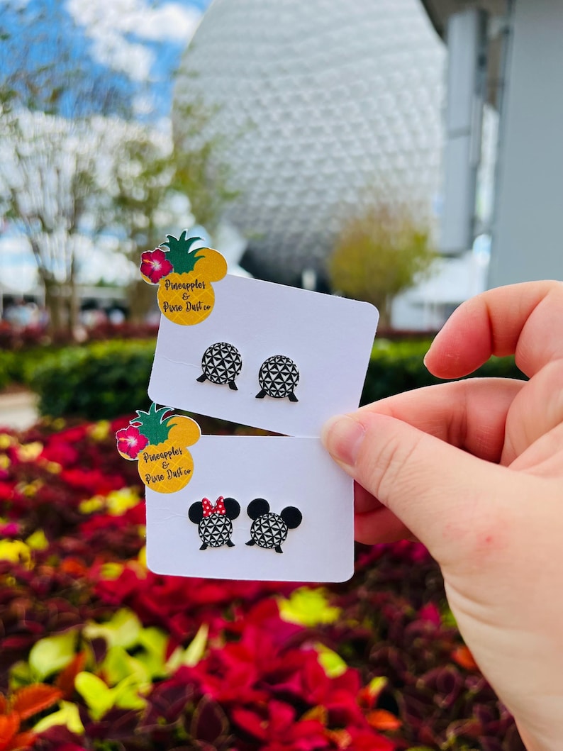 May include: Two pairs of stud earrings on white cards are held up in front of a blurred background. One pair features a geometric design, and the other has a Minnie Mouse design with a red bow. The cards have a pineapple logo with text.