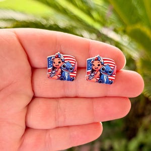 May include: A pair of square-shaped stud earrings featuring a cartoon illustration of a girl and a blue alien character, set against a backdrop of a red, white, and blue American flag design. The earrings are small and likely made of a glossy material.
