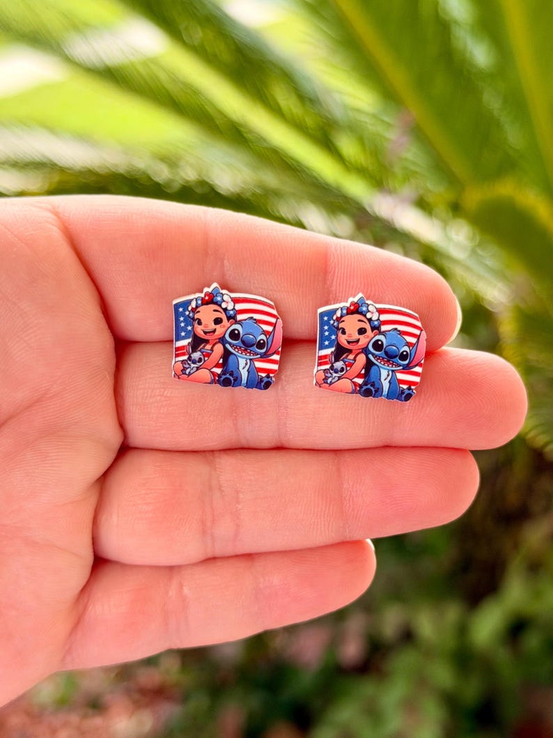 May include: A pair of square-shaped stud earrings featuring a cartoon illustration of a girl and a blue alien character, set against a backdrop of a red, white, and blue American flag design. The earrings are small and likely made of a glossy material.