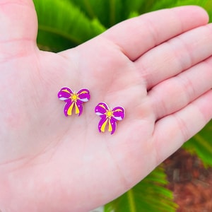 May include: Two small, purple and yellow bow-shaped earrings. The earrings have a bright yellow center and are held in an open hand against a blurred green background. The bows have a purple outline and are filled with a lighter purple color.