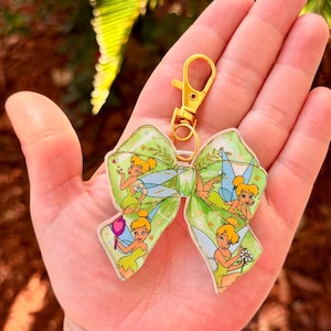 May include: A green bow-shaped keychain with a gold-colored clasp. The bow features illustrations of a fairy in a yellow dress with green wings. The background of the bow is a light green color with a leaf pattern.
