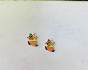 May include: Two cartoon mouse-shaped embellishments. Each mouse has a brown body, a light green shirt, and a green hat. The mice are in a running pose with their arms outstretched. The background is a white surface.