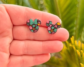May include: A pair of stud earrings featuring a Mickey Mouse silhouette design. The earrings have a black background with a colorful tropical floral pattern. One earring has a green bow.