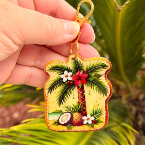 May include: A tropical-themed keychain featuring a palm tree, pineapple, and coconut design. The keychain has a gold-colored clasp and ring. The artwork includes red, white, and green accents on a yellow background.