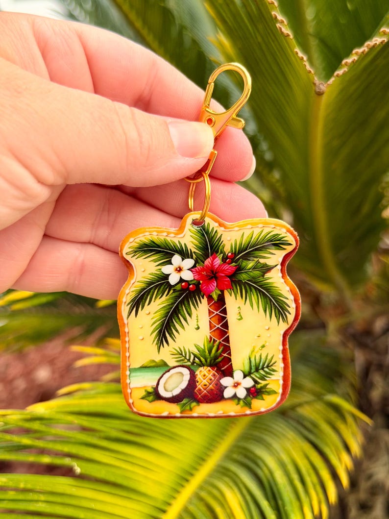 May include: A tropical-themed keychain featuring a palm tree, pineapple, and coconut design. The keychain has a gold-colored clasp and ring. The artwork includes red, white, and green accents on a yellow background.