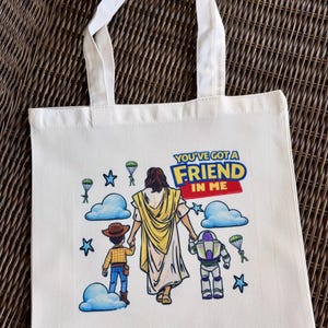 Pu&ograve; includere: Borsa tote in tela color crema con manici bianchi. La borsa presenta una grafica colorata di Ges&ugrave; che tiene per mano Woody e Buzz Lightyear, con la scritta "YOU'VE GOT A FRIEND IN ME". Nuvole blu, stelle e paracadute completano il design.