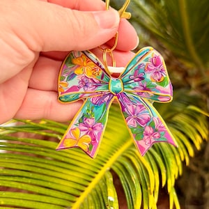 May include: A colorful bow-shaped keychain with a gold-colored clasp. The bow features a floral design with pink, yellow, and blue flowers on a teal background. The keychain is held against a backdrop of green palm leaves.