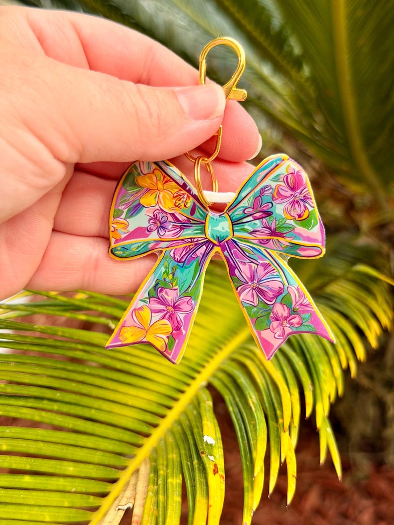 May include: A colorful bow-shaped keychain with a gold-colored clasp. The bow features a floral design with pink, yellow, and blue flowers on a teal background. The keychain is held against a backdrop of green palm leaves.