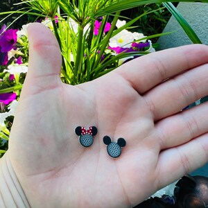May include: Two small, black and silver stud earrings shaped like Mickey Mouse heads. One earring features a red and white polka dot bow. The earrings are displayed on a light skin tone hand.