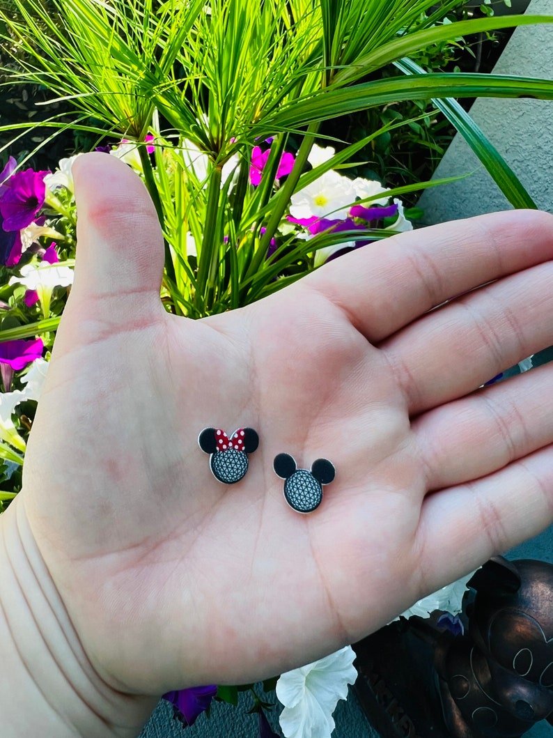 May include: Two small, black and silver stud earrings shaped like Mickey Mouse heads. One earring features a red and white polka dot bow. The earrings are displayed on a light skin tone hand.