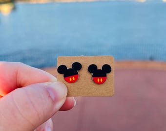 May include: A pair of black and red Mickey Mouse stud earrings. Each earring features a black head with red shorts and yellow accents. The earrings are displayed on a small, brown cardboard backing.