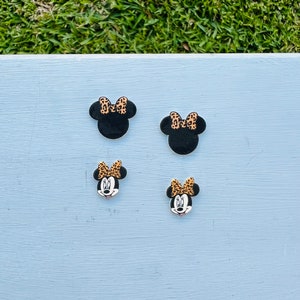 May include: Four black Minnie Mouse-shaped embellishments with leopard print bows. Two are simple silhouettes, and two feature Minnie's face. The embellishments are arranged on a light blue surface.
