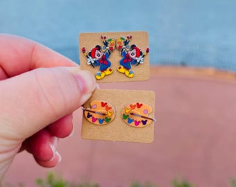 Op de afbeelding: Twee paar emaille oorbellen. Eén paar heeft een stripfiguur in een schildersoutfit met penselen. Het andere paar is een paletontwerp met kleurrijke Mickey Mouse-hoofden. Beide paren zitten op kleine bruine kaarten.