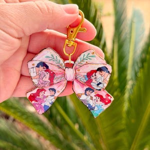 May include: Pink bow-shaped keychain featuring a cartoon image of a red-haired woman and a man in a blue jacket. The bow has a gold-colored clasp. The background is blurred green.