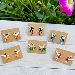 May include: A collection of six pairs of fairy-themed stud earrings. Each pair features a different fairy design with colorful outfits and white wings. The earrings are displayed on small, rectangular brown cards.